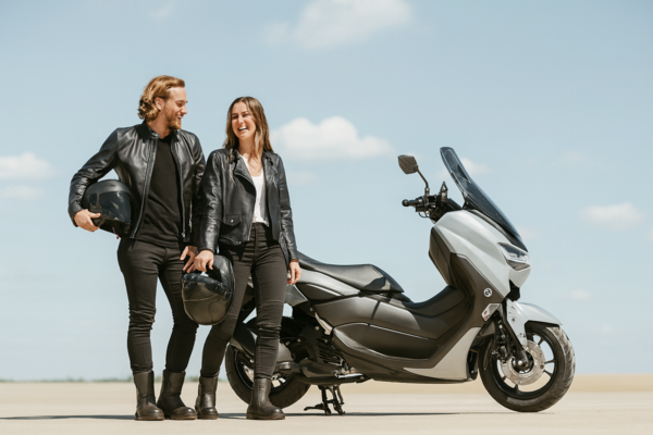 Junges Paar in schwarzer Motorradkleidung steht lachend neben einem silbernen Yamaha NMAX Roller, beide mit Helm in der Hand – Fahrschule Lind Berlin B196 Ausbildung.