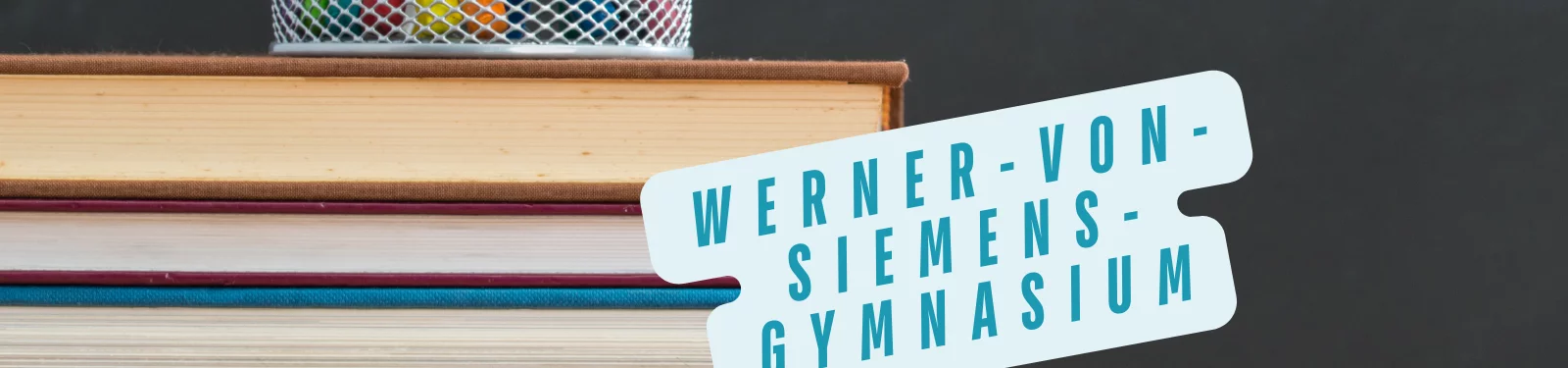 Ein Stapel Bücher mit einem Stiftehalter auf einem Tisch. Auf einem blauen Etikett steht 'Werner-von-Siemens-Gymnasium'