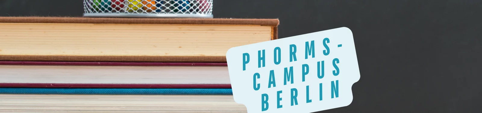 Ein Stapel Bücher mit einem Stiftehalter obenauf, neben einem Schild mit der Aufschrift 'Phorms-Campus Berlin'.
