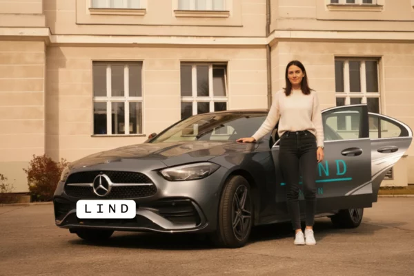 Junge Frau steht neben einem Mercedes-Fahrschulfahrzeug der Fahrschule Lind in Berlin vor einem Gebäude. geöffnete Autotür, modernes Ausbildungsfahrzeug.
