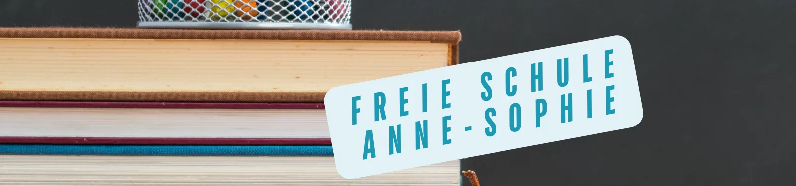 Ein Stapel Bücher mit einem Stiftehalter im Hintergrund. Vor dem Stapel ist ein schräg platzierter Schriftzug zu sehen, der 'Freie Schule Anne-Sophie' in blauen Großbuchstaben zeigt