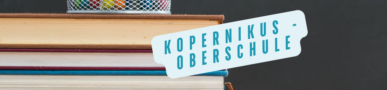 Das Bild zeigt einen Stapel mehrerer Bücher in verschiedenen Farben. Oben auf den Büchern steht ein kleiner runder Metallkorb, der mit bunten Stiften gefüllt ist. Der Hintergrund ist dunkel und schlicht, was den Fokus auf den Büchern und dem Korb lässt. Über den Büchern befindet sich ein schräg platzierter Text in einer hellblauen Blase, auf der in großen, klaren Buchstaben „Kopernikus-Oberschule“ steht.