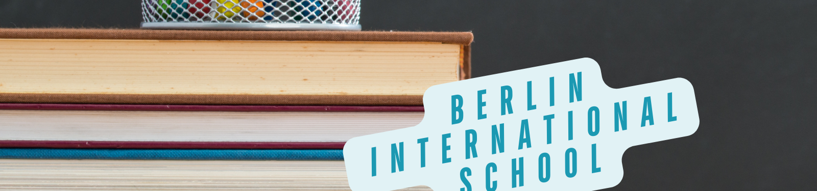 Ein Stapel Bücher in verschiedenen Farben mit einem Stiftehalter voller bunter Stifte auf dem obersten Buch. Im Vordergrund befindet sich ein Schild mit der Aufschrift "Berlin International School" in blauen Großbuchstaben auf einem weißen Hintergrund. Das Bild vermittelt ein Gefühl von Lernen und Bildung an der internationalen Schule in Berlin.