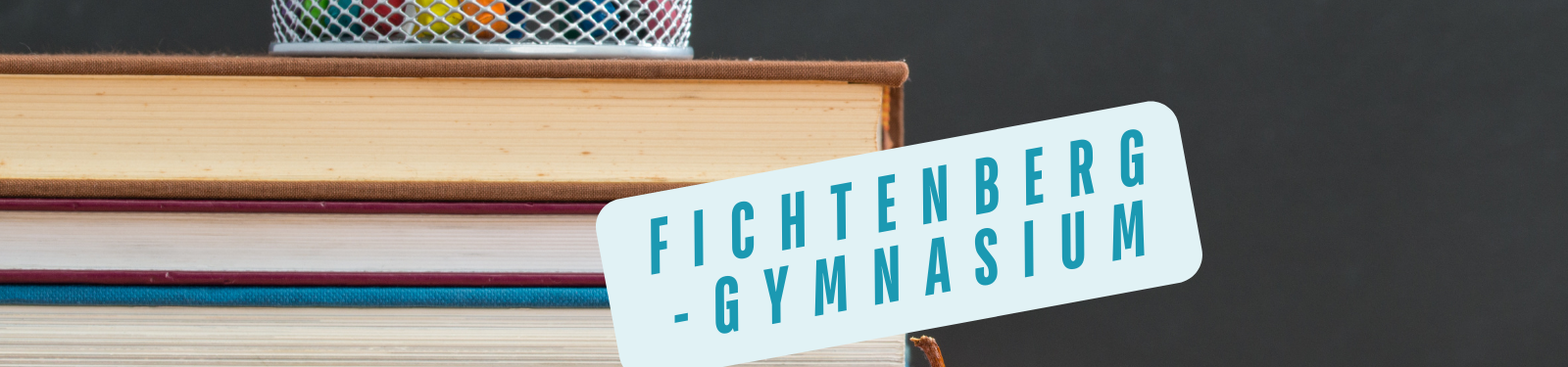 Ein Bild, das Bücher in verschiedenen Farben zeigt, gestapelt mit einem Stifthalter oben drauf. Ein Schild daneben trägt den Schriftzug Fichtenberg-Gymnasium