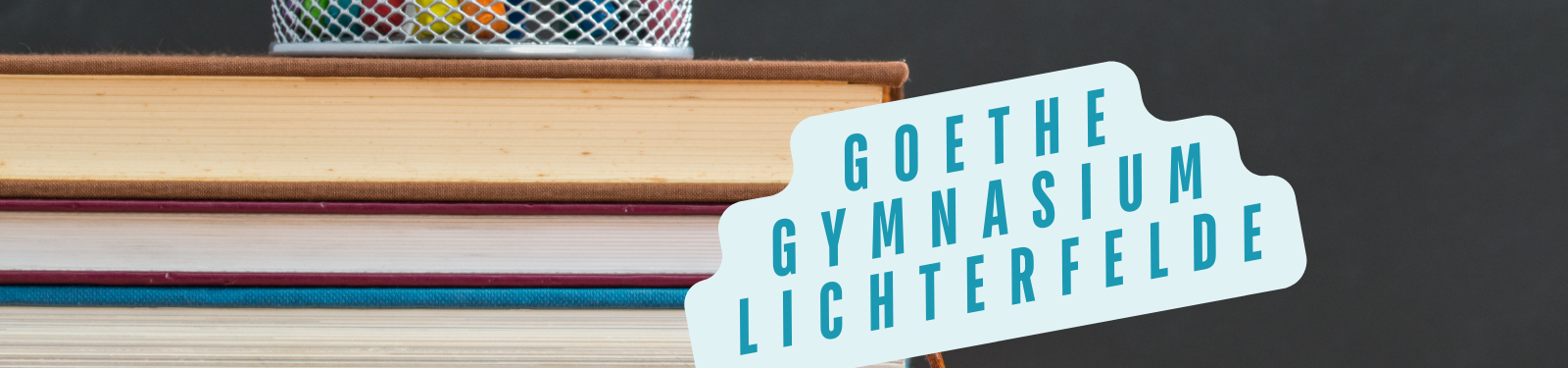 Ein Stapel Bücher mit einem Stiftehalter darauf. Ein Schild zeigt den Text "Goethe Gymnasium Lichterfelde"