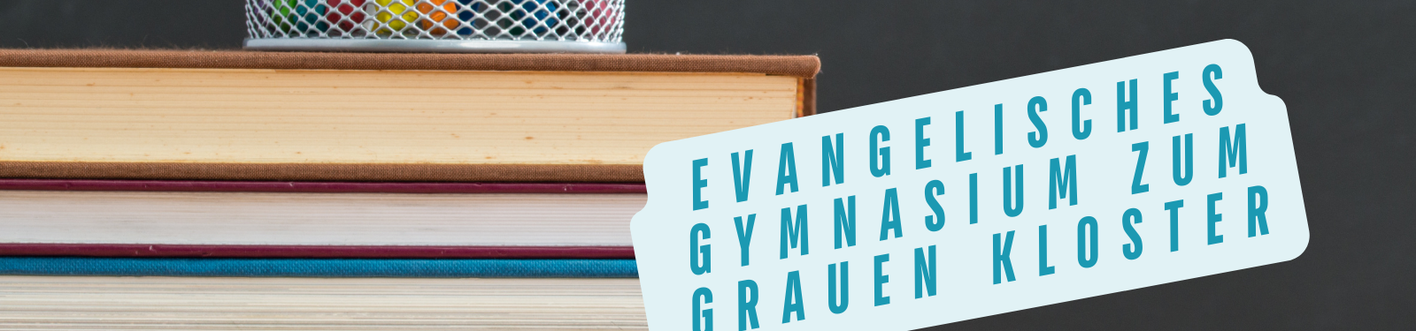 Stapel Bücher mit einem Stiftehalter darauf, daneben ein Schild mit der Aufschrift 'Evangelisches Gymnasium zum Grauen Kloster