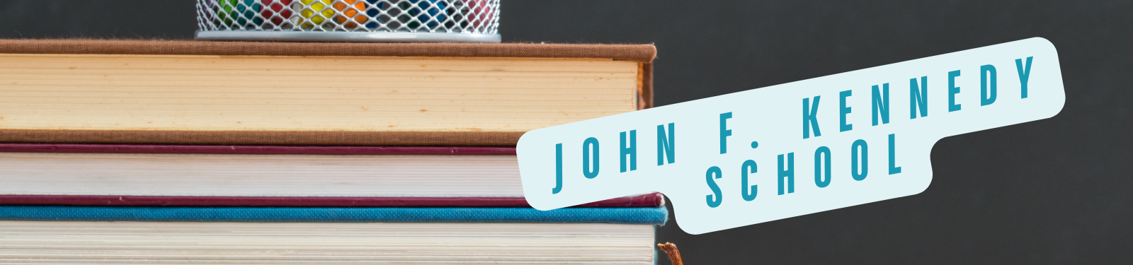 Ein Stapel Bücher mit einem Stiftehalter obenauf. Ein hellblaues Banner mit der Aufschrift "John F. Kennedy School" befindet sich im Vordergrund.