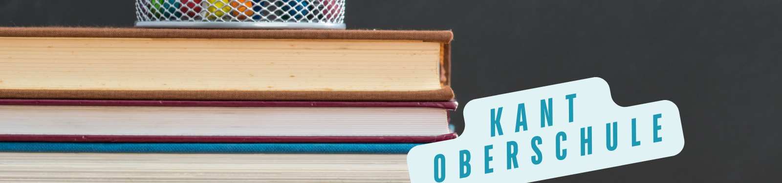 Ein Stapel Bücher mit einem Stiftehalter obenauf, daneben ein Schild mit der Aufschrift "Kant Oberschule" in blauen Buchstaben.