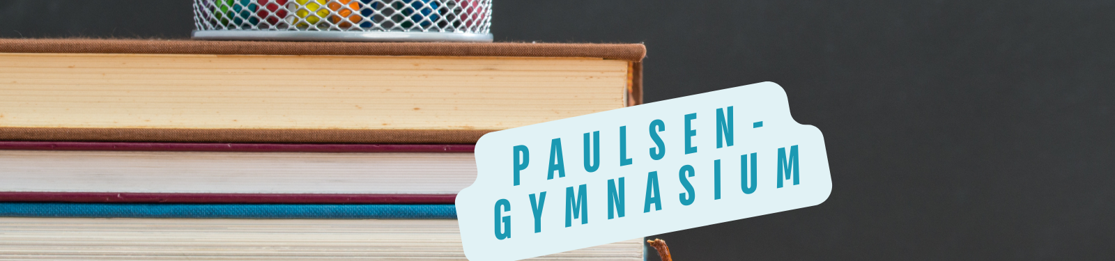 Ein Stapel Bücher mit einem Stiftehalter oben drauf, der bunte Stifte enthält. Ein weißes Schild mit blauer Aufschrift "Paulsen-Gymnasium" ist an der Seite des Bücherstapels angebracht. Der Hintergrund ist dunkel und hebt die hellen Farben des Buchrückens und des Schildes hervor.