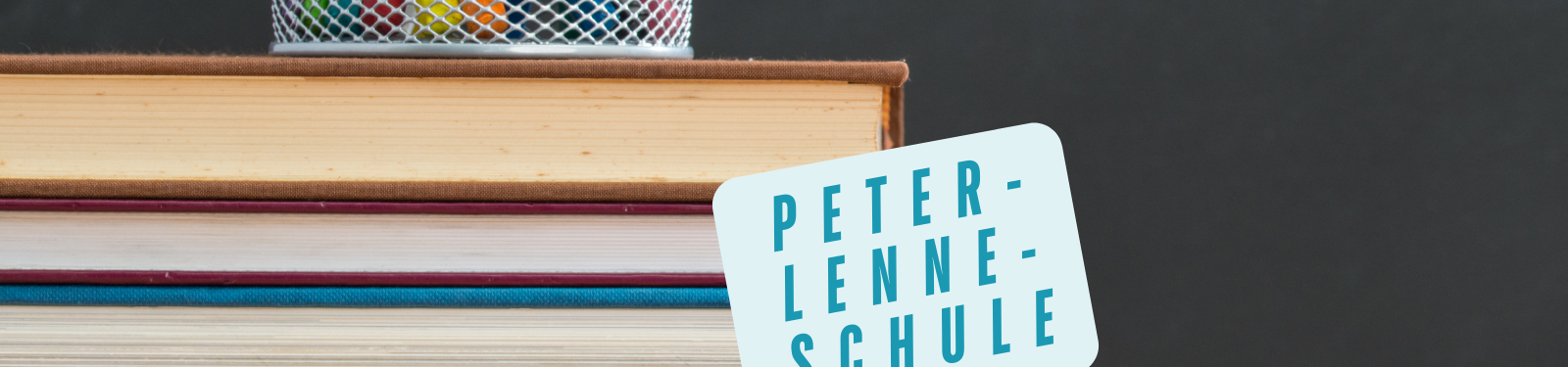 Ein Stapel Bücher mit einem Stiftehalter obenauf. Auf einem der Bücher ist ein Schild mit der Aufschrift 'Peter-Lenne-Schule' angebracht.