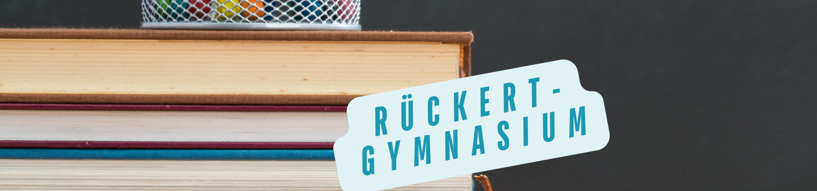 Ein Stapel Bücher mit einem Stiftehalter darauf und einem Schild mit der Aufschrift 'Rückert-Gymnasium'
