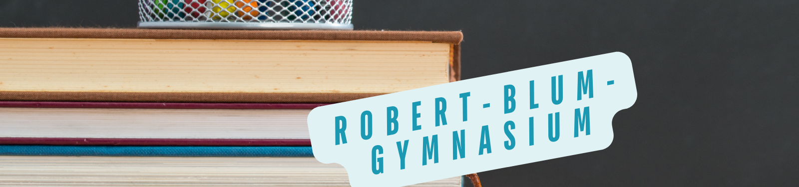 Ein Stapel Bücher, auf denen ein Schild mit der Aufschrift "Robert-Blum-Gymnasium" liegt. Im Hintergrund steht ein Stiftehalter mit bunten Stiften.