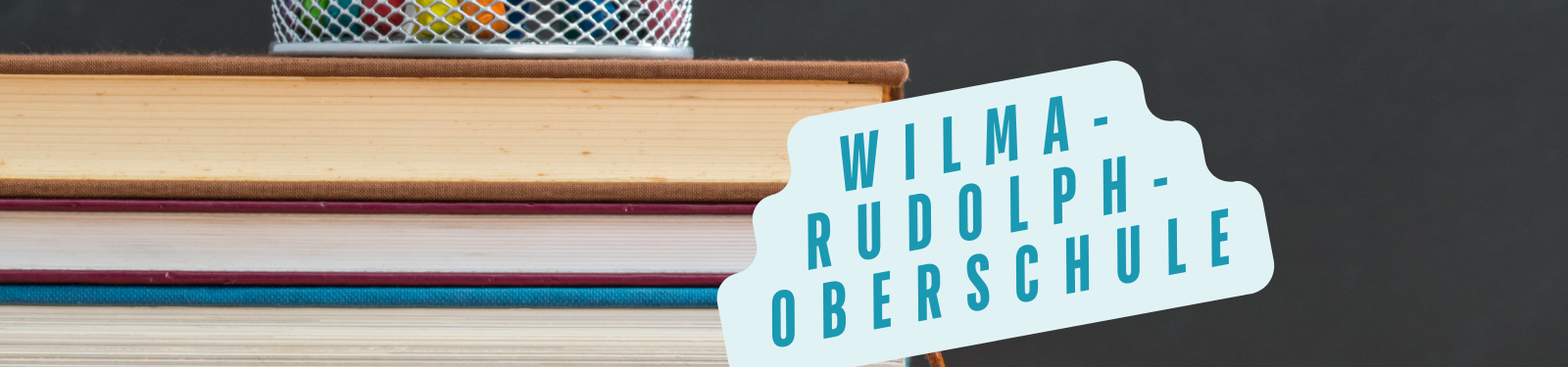 Ein Stapel Bücher mit einem Stiftehalter darauf. Ein Schild zeigt den Text Wilma-Rudolph-Oberschule